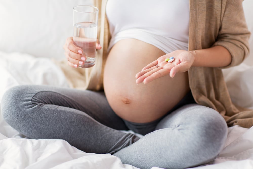 Une femme enceinte tenant un verre d'eau et des suppléments des laboratoires Hollis.