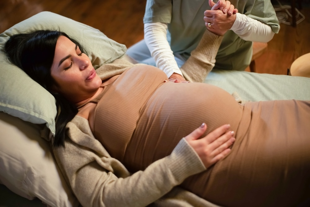 Une femme enceinte vit la phase de latence de son accouchement, allongée sur un lit, les mains sur le ventre.