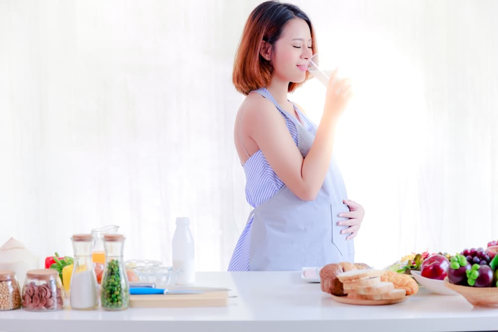 Une femme enceinte en tablier, affectueusement surnommée « maman », se tient gracieusement devant une cuisine. Elle tient un verre d'élixir doré, lait d'or
