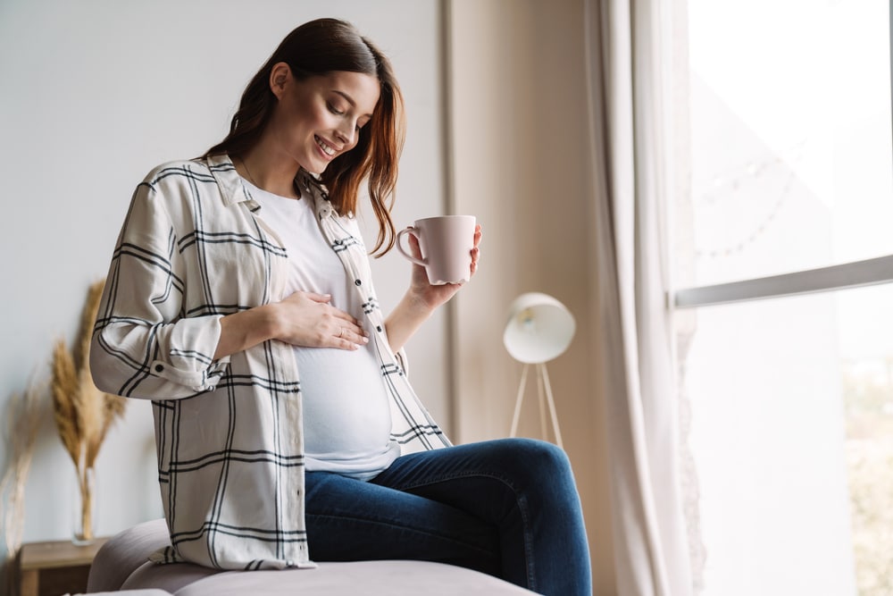 Une femme enceinte assise sur un lit tenant une tasse de café.
