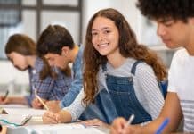 Une jeune femme souriante regardant la caméra tout en écrivant dans un cahier dans une salle de classe avec d'autres élèves concentrés sur leur travail, symbolisant la réussite scolaire.