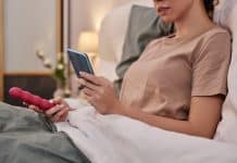 Une personne est allongée dans son lit, tenant un smartphone dans une main et un Sextoy dans l'autre.