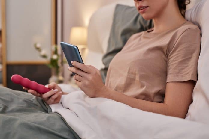 Une personne est allongée dans son lit, tenant un smartphone dans une main et un Sextoy dans l'autre.