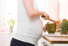 Une femme enceinte vêtue d'un t-shirt blanc et d'un short bleu marine coupant une ananas sur un comptoir de cuisine.
