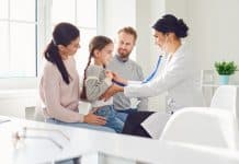 Un médecin examine une jeune fille avec un stéthoscope dans une clinique, tandis que ses parents la regardent avec des sourires affectueux, considérant les critères complémentaires de santé pour leur famille.