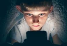Un jeune garçon sous une couverture, éclairé par un écran lumineux, regardant intensément un smartphone dans une pièce sombre, inconscient de l'importance de la protection des données.