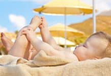 Un bébé allongé sur une serviette sous le soleil, se tenant les pieds de manière ludique avec des parasols et un ciel dégagé en arrière-plan.