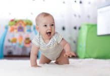 Un bébé avec un sourire à bouche ouverte rampe sur un tapis blanc dans une salle de jeux, entouré de jouets colorés, illustrant son développement préco