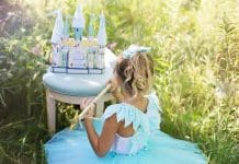 Une jeune fille vêtue d'une robe de princesse est assise à l'extérieur et joue avec un château jouet sur un tabouret, entouré d'herbes hautes.