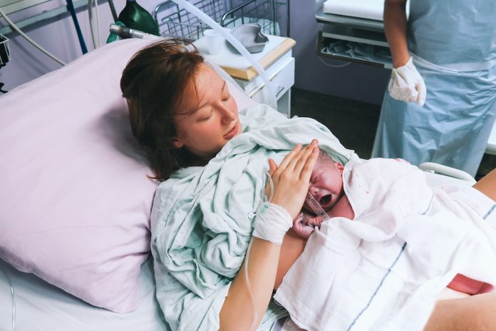Une femme est allongée dans un lit d'hôpital, tenant un nouveau-né enveloppé dans une couverture blanche sur sa poitrine, réfléchissant aux différentes phases de l'accouchement. Une autre personne portant des gants se tient à proximité.